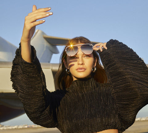 A female model standing in front of a plane wearing a black top with QUAY HIGH KEY aviator sunnies