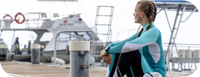 Person in wetsuit sitting on a dock, looking at the water.