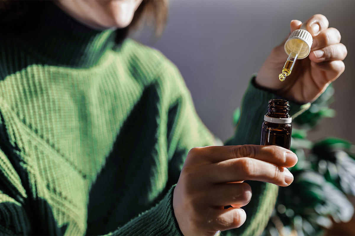 A woman in a green sweater carefully holds a dropper with golden CBD oil over a small amber glass bottle. Sunlight highlights her hands and the natural product.