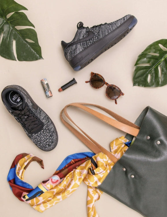 The summer style edit flat lay shows the Nike Fly Knit running shoe in charcoal gray, with a matching tote purse. Its contents are spilling out revealing a brightly printed silk scarf, brown sunglasses, and red lipstick. 