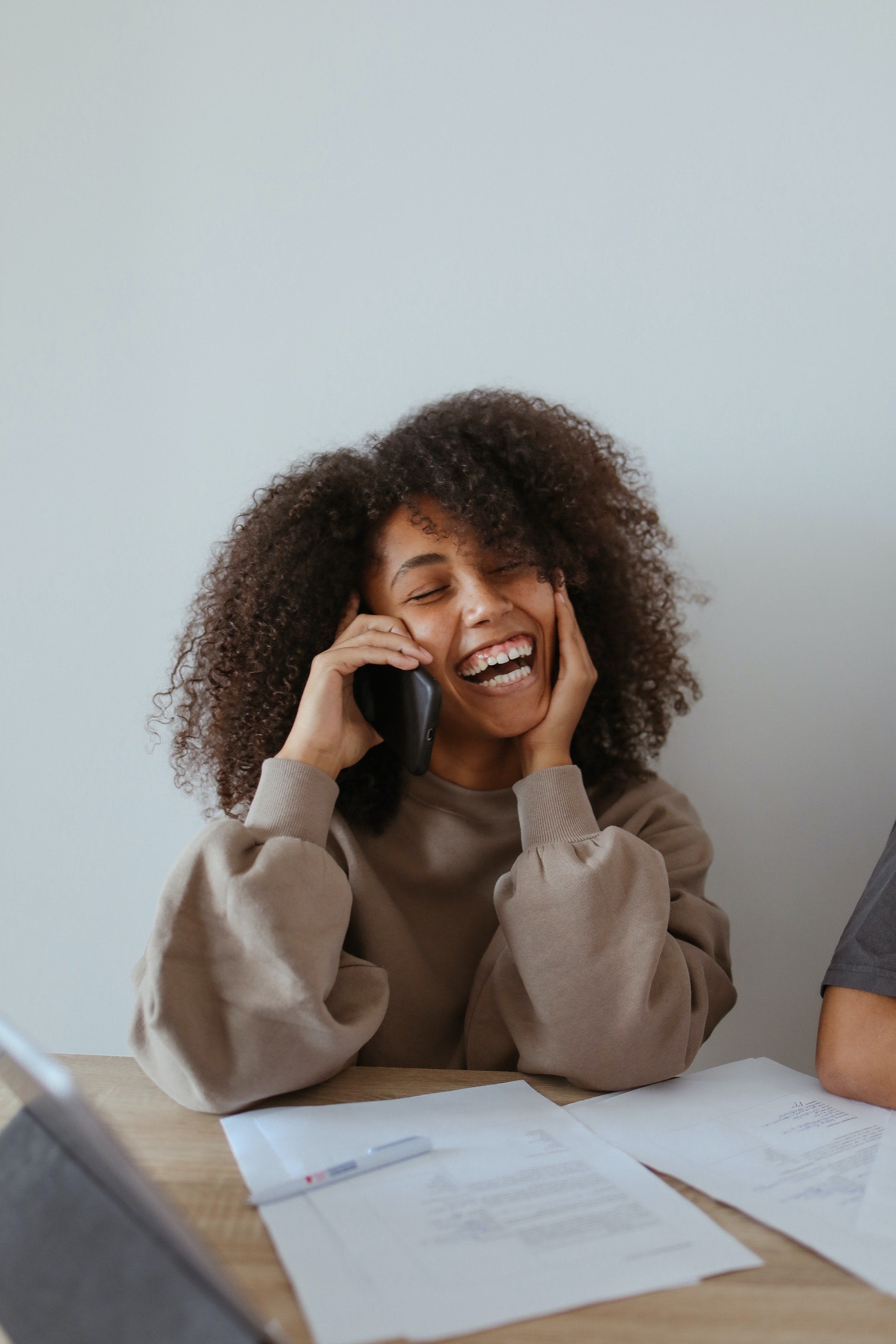 Woman sitting at a desk laughing. She has brown curly hair and is wearing a brown sweater. The desk has papers in front of her.