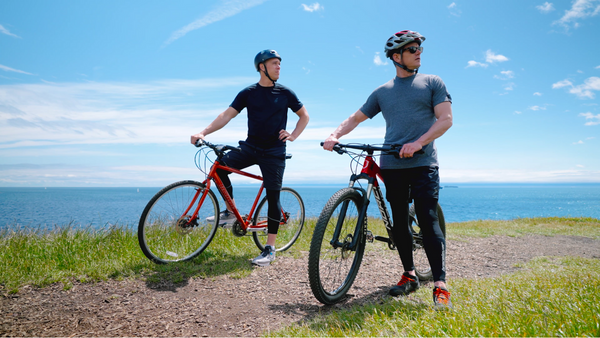 Two men on bicycles, on a cliff with the sea behind them. They are wearing Bracelayer's knee support cycling pants