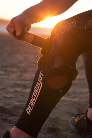On a beach at sunset, Bracelayer® founder and CEO Dan Tognotti straps a custom fit knee brace on over his Bracelayer® compression pants with built-in knee support, designed to help keep such braces locked in their places.