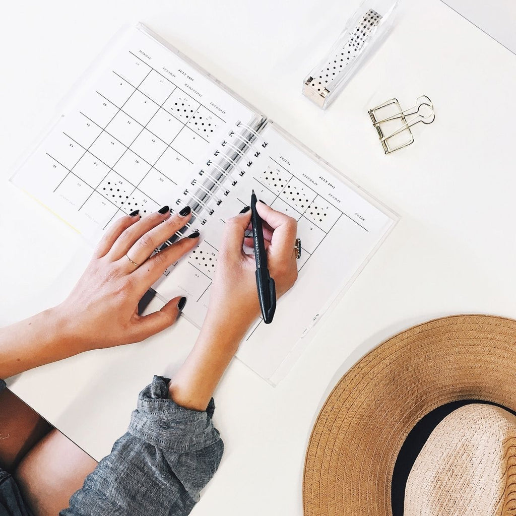 Ladies hands with black painted nails and straw hat on the table writing notes in an weekly planning book