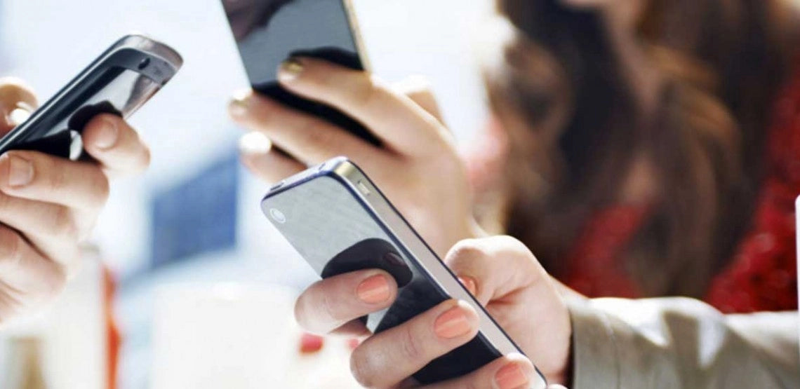 A close-up of several people’s hands holding smartphones, each person focused on their device, with a blurred background showing a woman in a red outfit.