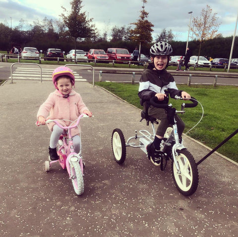 Image of a young girl and boy riding on their bikes side by side outside. The boys bicycle is an adapted bike for children with special needs