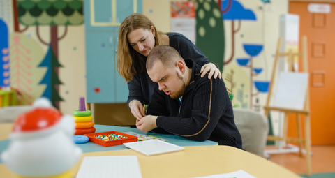 a young adult with learning disabilities in a learning environment. A female teacher is leaning over, observing him.