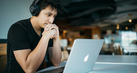 hombre sentado en una oficina mirando la pantalla de una computadora portátil. Con los codos sobre la mesa y las manos en la cara, usando auriculares.