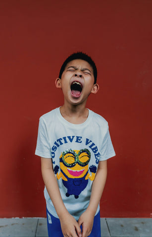 young boy against a red background. His eyes are closed and his mouth is wide open as though he is screaming.