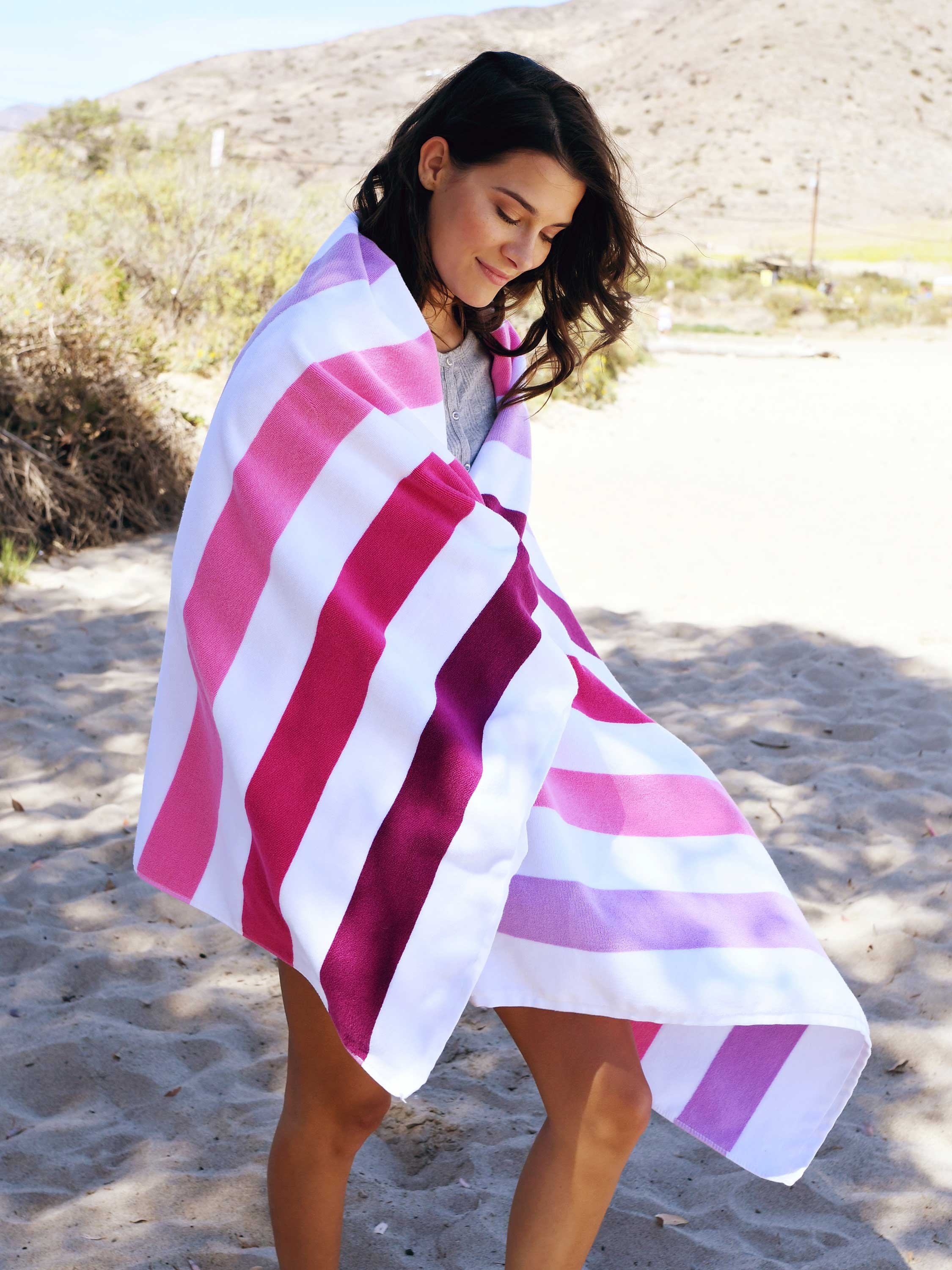 A woman walking on the beach draped in a pink striped Cabana Towel.