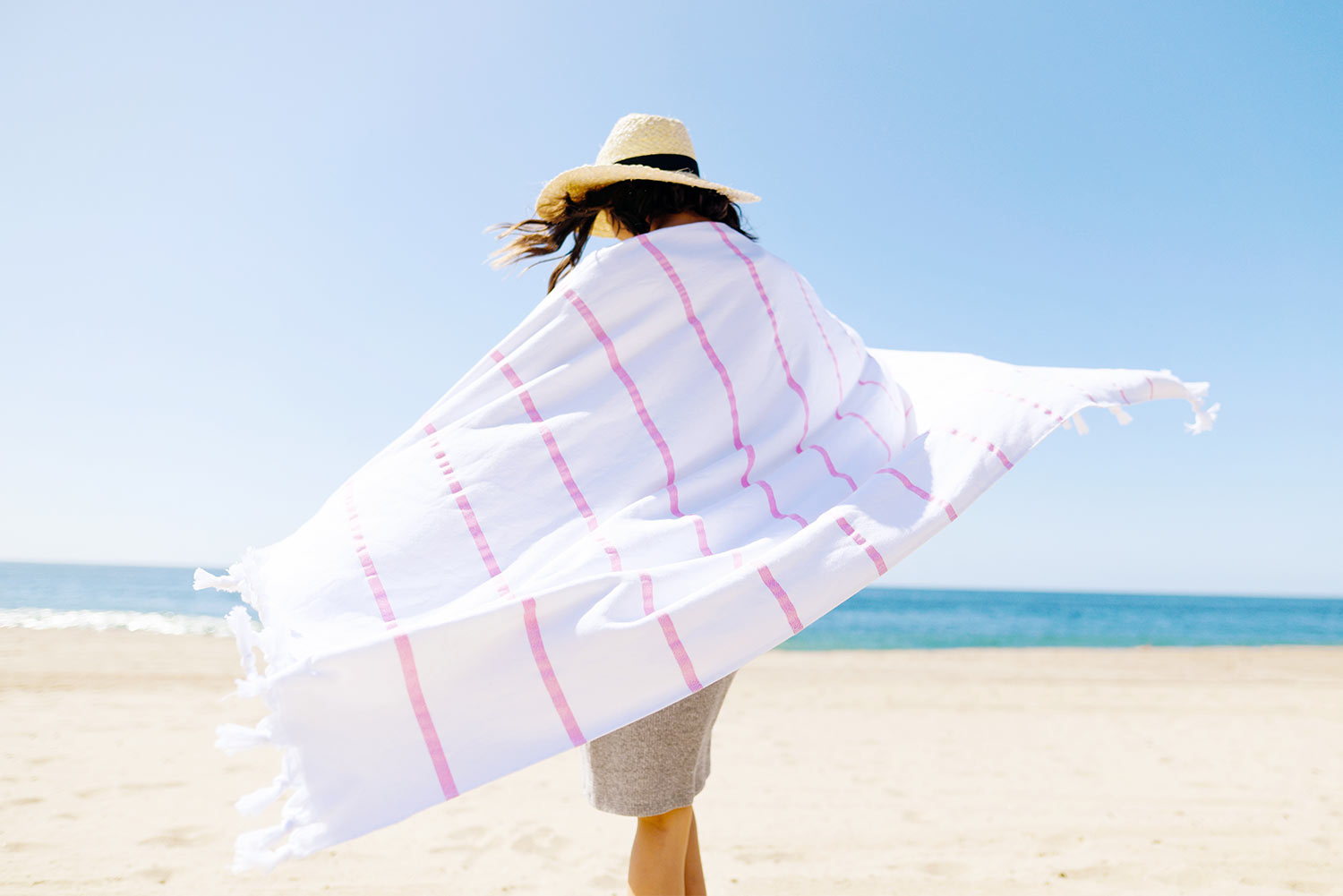 A woman walking towards the ocean draped in a pink striped Turkish Towel.