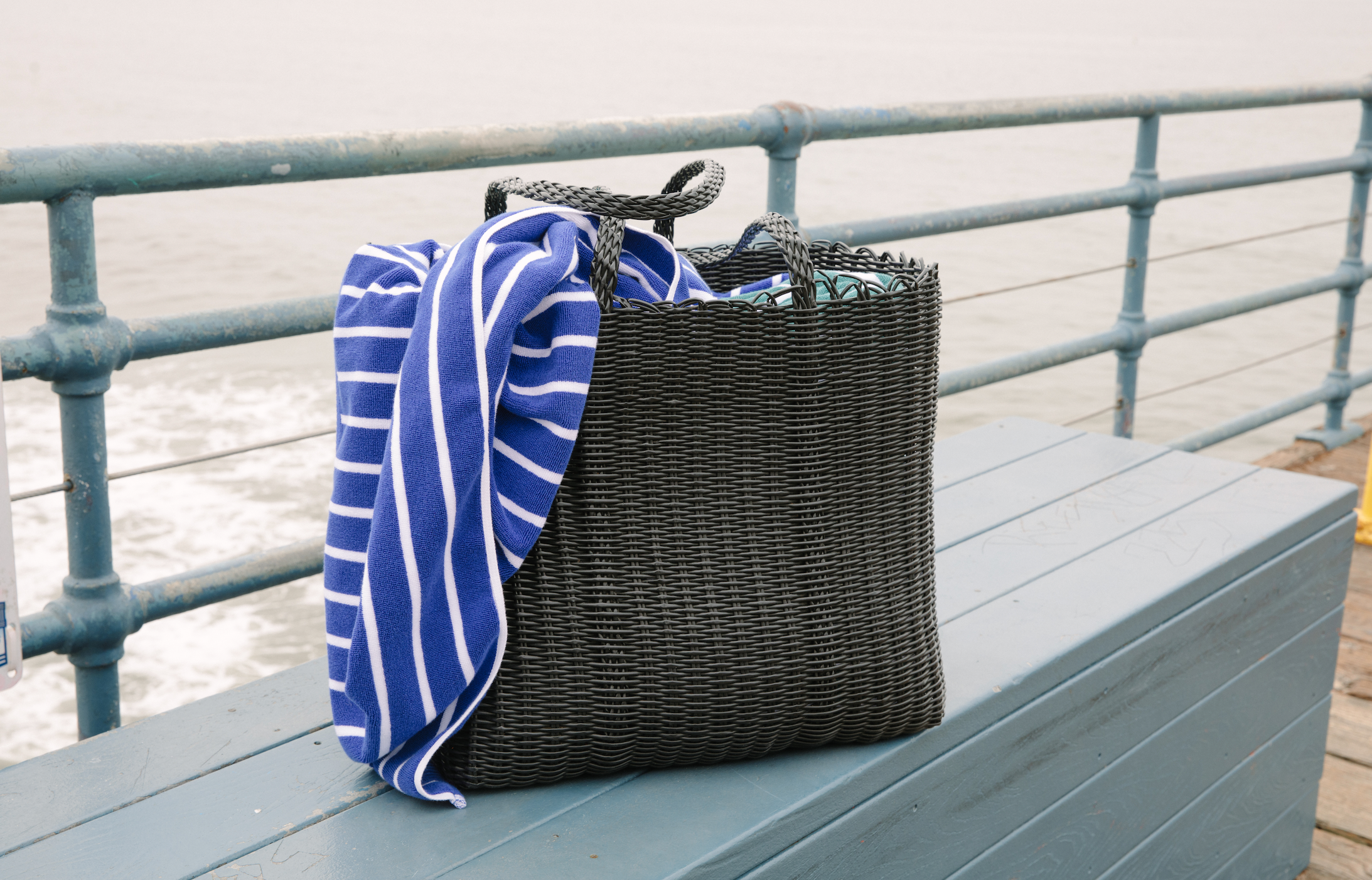 A black woven beach bag with a Cabana towel draped inside.