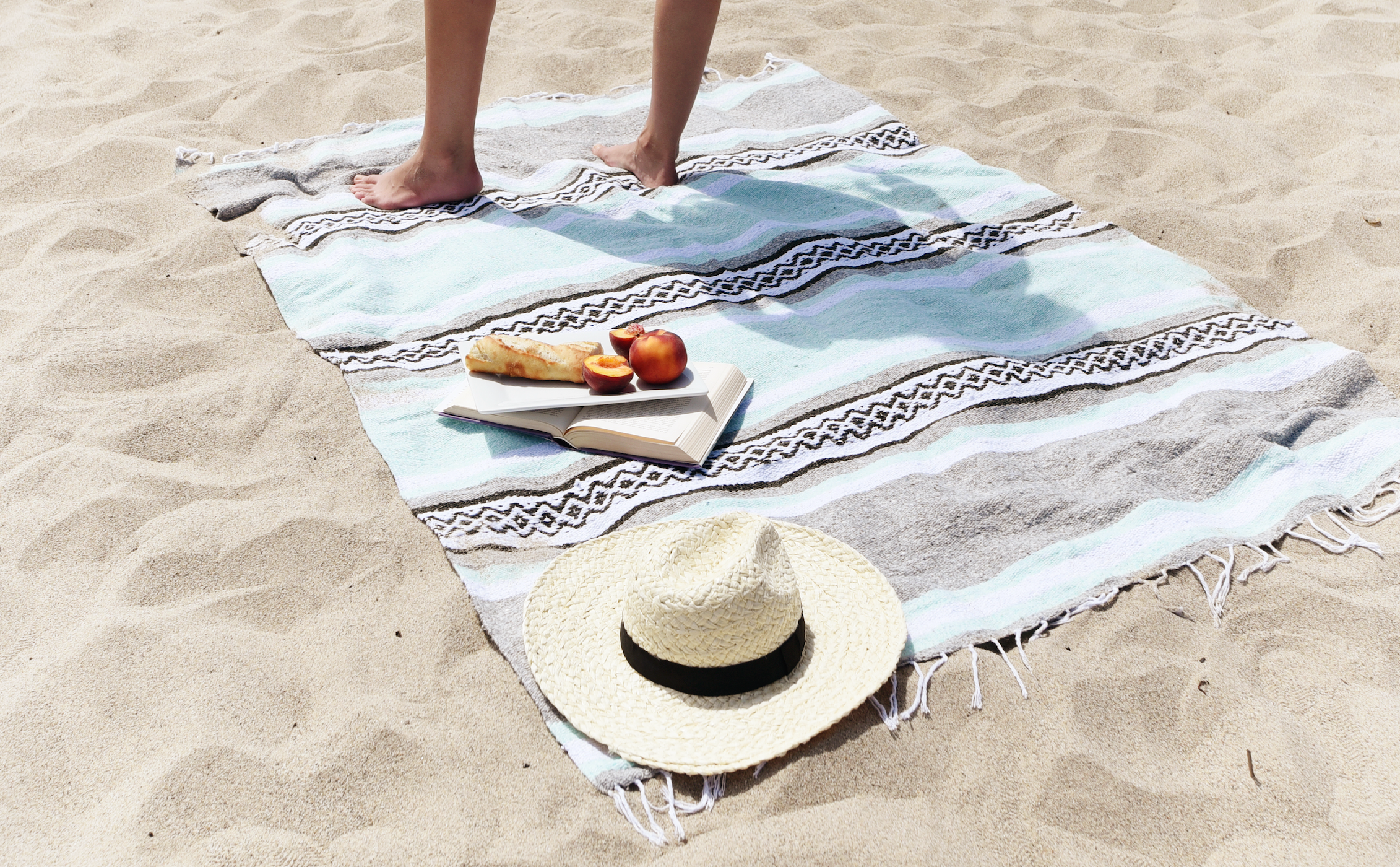 A Mexican blanket laid on the sand with a hat, book, and a woman's leg standing on top.