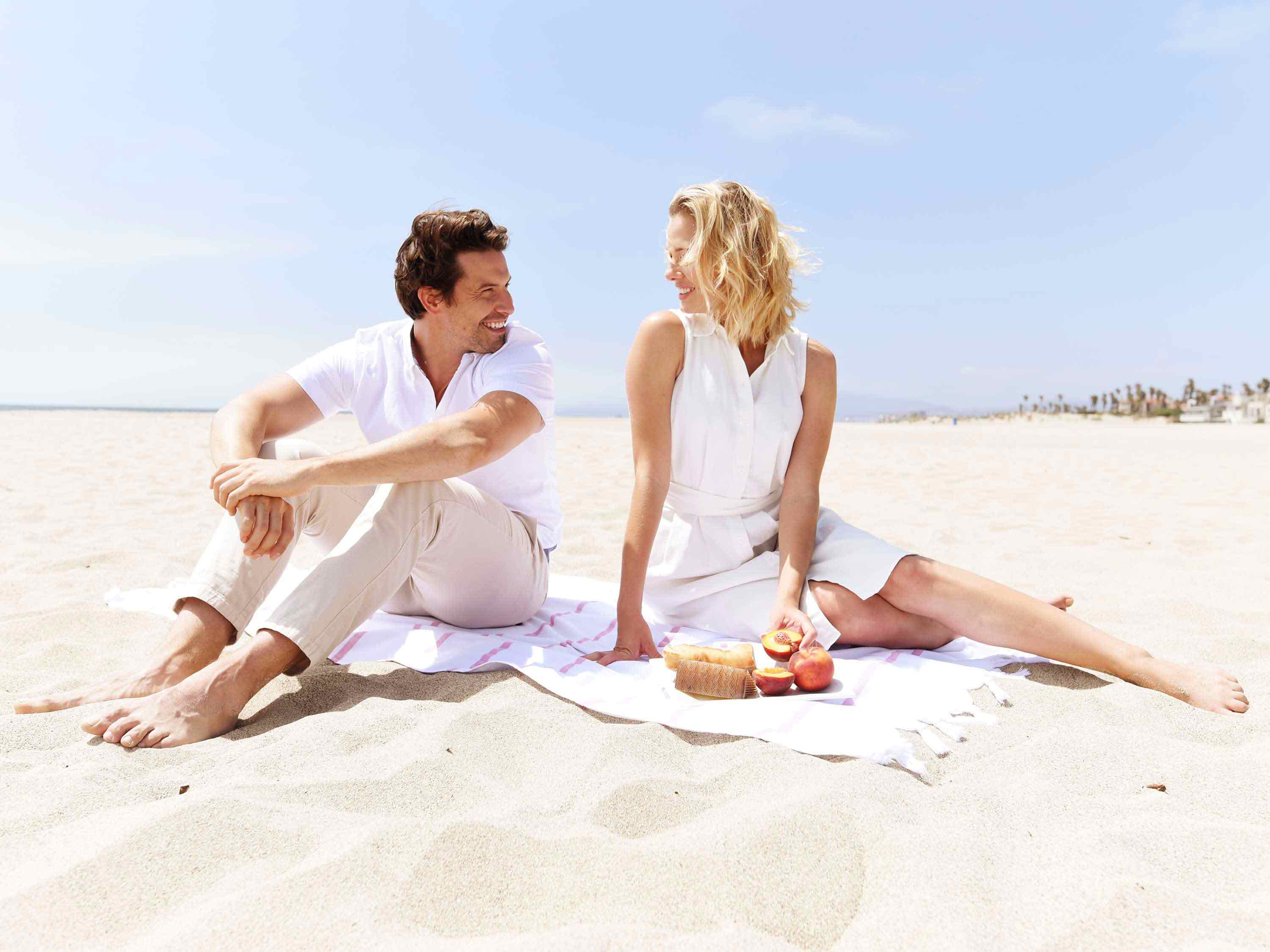 A couple sitting on a Turkish Towel looking at each other on the beach.