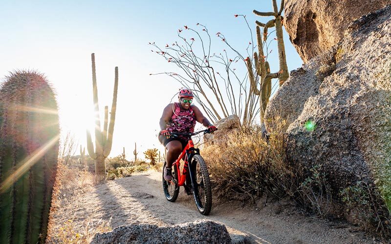 Mirna rides her Timberjack past a large rock formation on an Arizona trail, surrounded by cacti of all sorts.