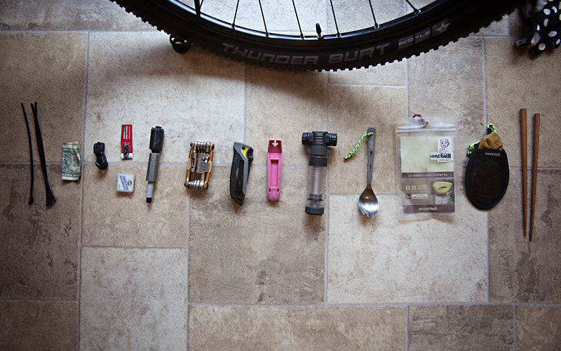 Gear and tools for bikepackging are lined up on the floor. The items include zip-ties, cash, a multi-tool, tire lever, spork, and chopsticks.