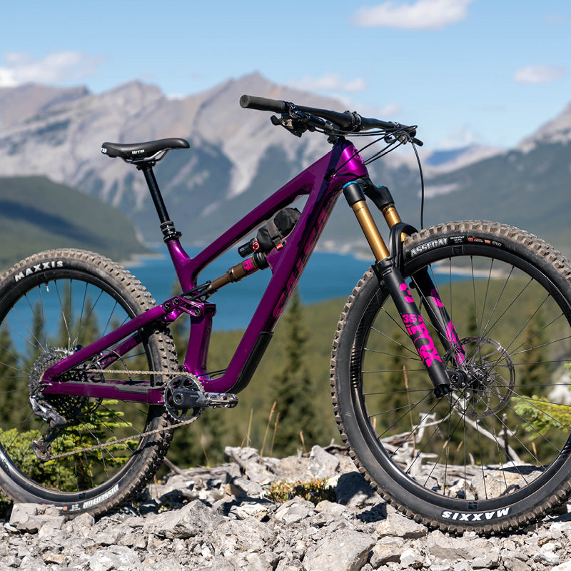 A vivid purple Salsa Blackthorn full-suspension mountain bike stands high in the Rocky Mountains above an alpine lake.