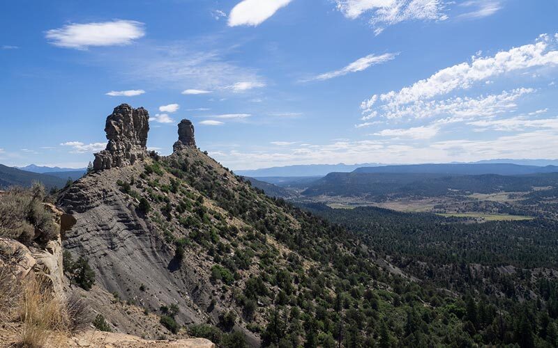 The twin sandstone spires of Chimney Rock National Monument stand under blue skies atop steep sparsely vegetated slopes.