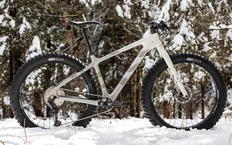 Beargrease Carbon SLX fat tire bike in white with gray graphics, stands in a snow-covered pine forest