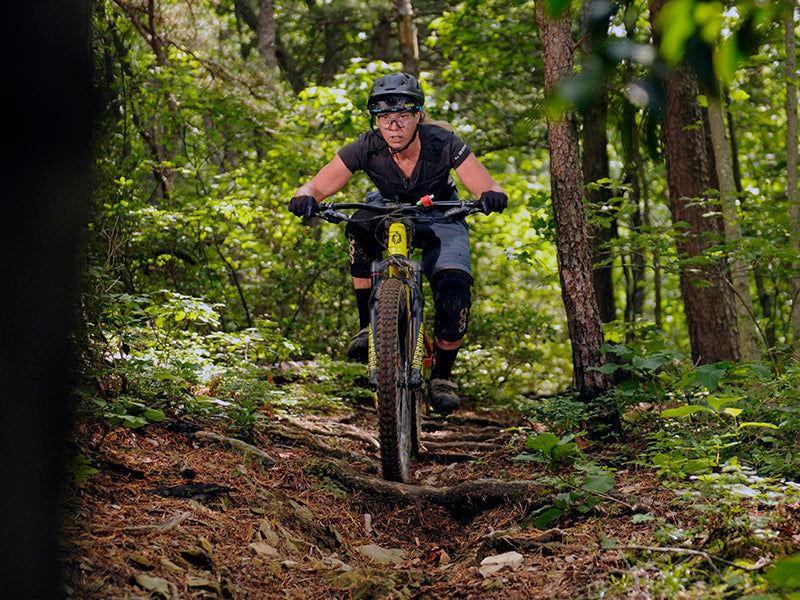 A rider rolling over a rooty section of singletrack in a green, wooded area.