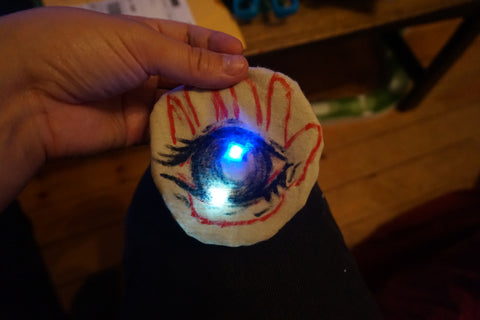 round patch with an black outline of an eye and a red hand outline behind it with the blue and white LEDs lit up