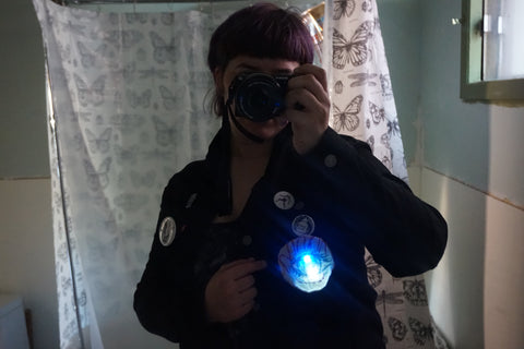 white person with purple hair standing in front of a mirror with the camera over their face, they are wearing a black denim jacket with a light up patch pinned to it