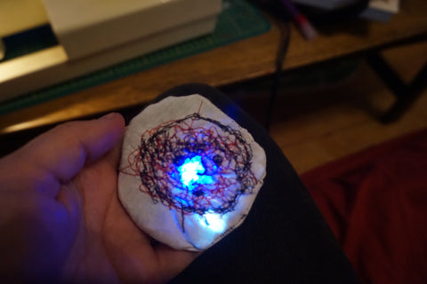  embroidered outline of a face in black thread over top of a chaotic mess of red thread with both the white and blue LEDs lit up