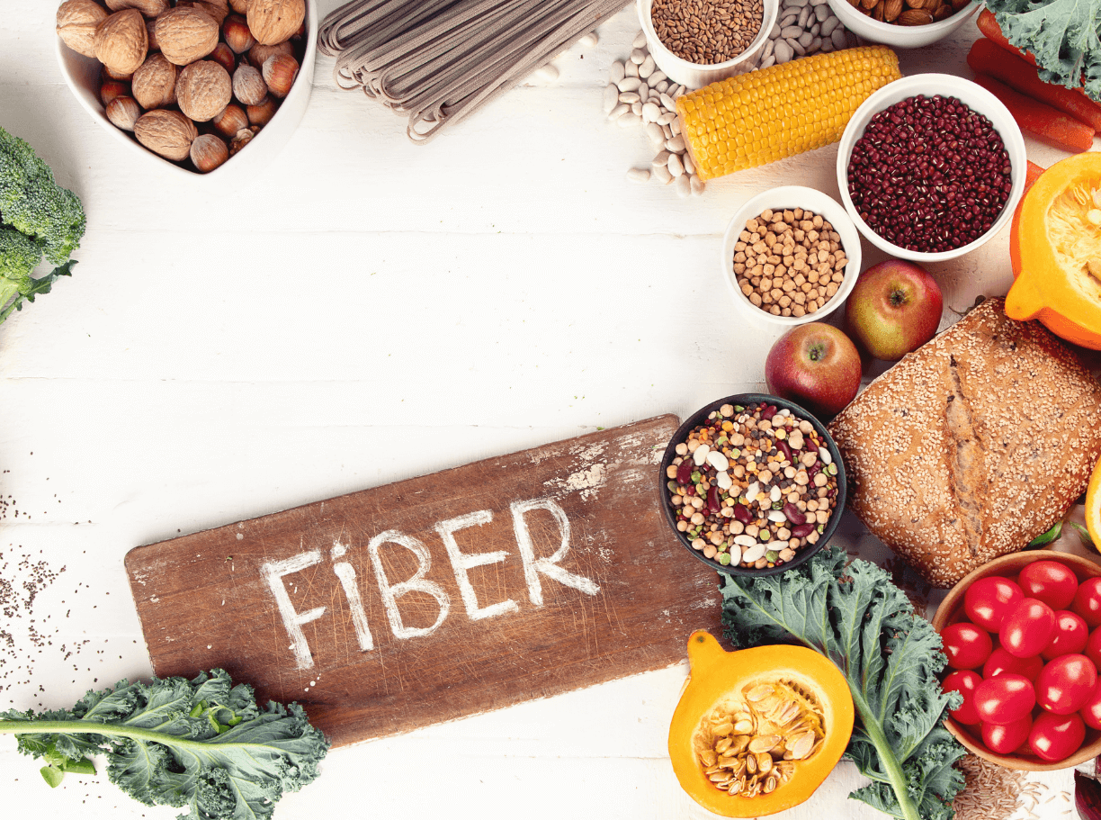 Alimentos ricos en fibra como frutas, verduras, legumbres, cereales integrales, frutos secos y pan integral, junto a cartel con la palabra fiber