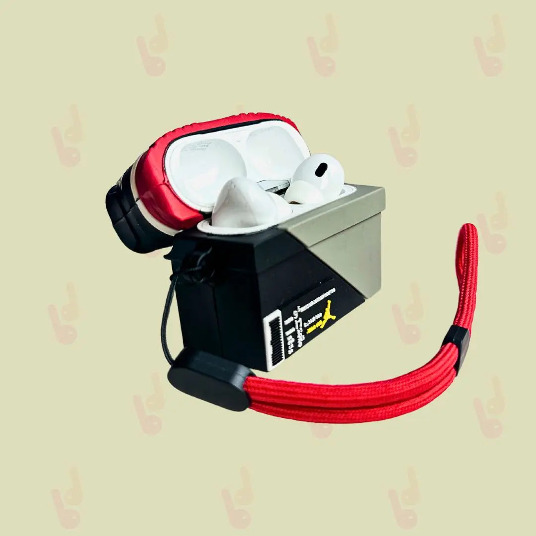 Side view of the Dawbaw Sneaker Snap AirPods case with the red sneaker lid open, showing earbuds inside the shoebox base, and a red lanyard attached.