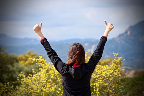 Picture of a woman overlooking a picturesque valley, with arms outstretched above her, with her thumbs up, donating victory, happiness, achievement