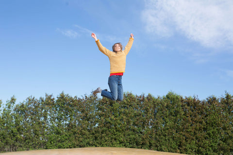 Woman jumping in the air with joy - a large blue sky is above her. She is dressed in yellow top and blue jeans