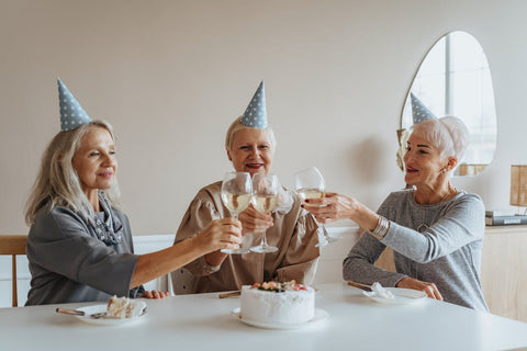 Mental Fitness - 3 older women sitting around a cake, wearing birthday hats, and cheersing glasses of white wine