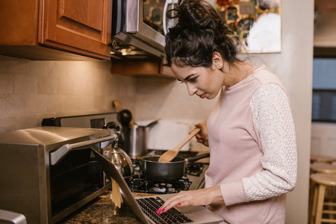 Mental Fitness - Woman in kitchen, looking at laptop while stirring a pot