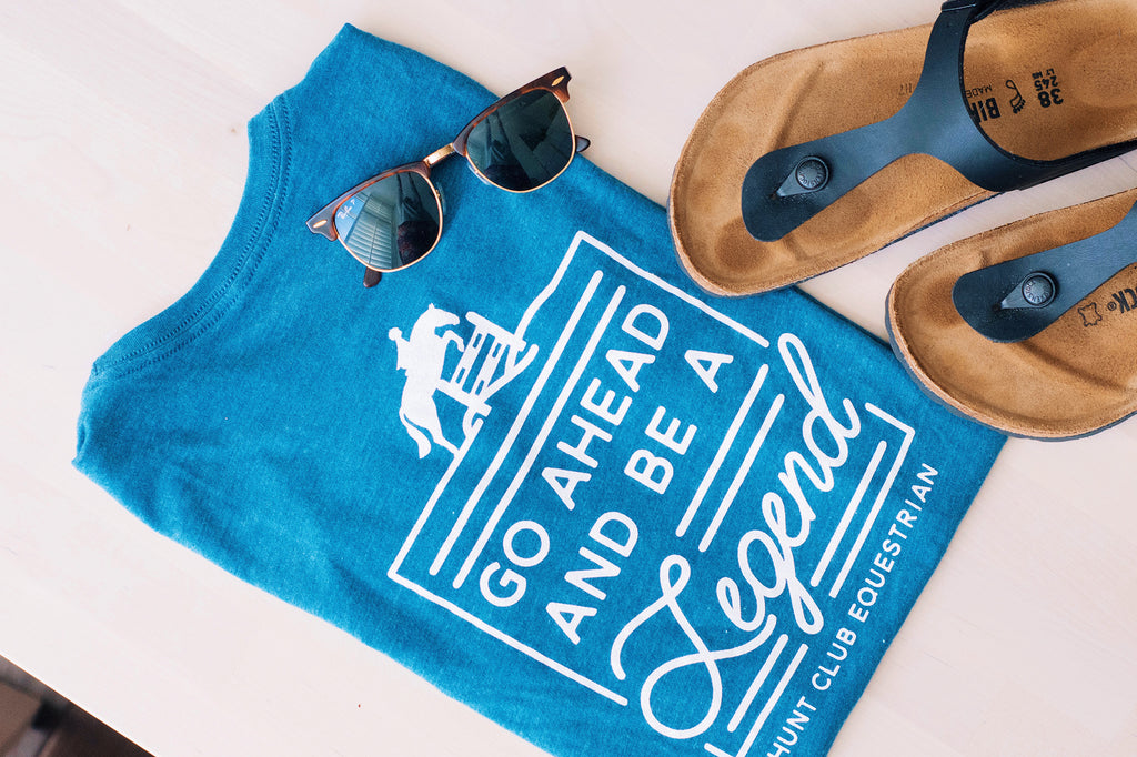 A teal Hunt Club tee next to a pair of sandals and sunglasses. 