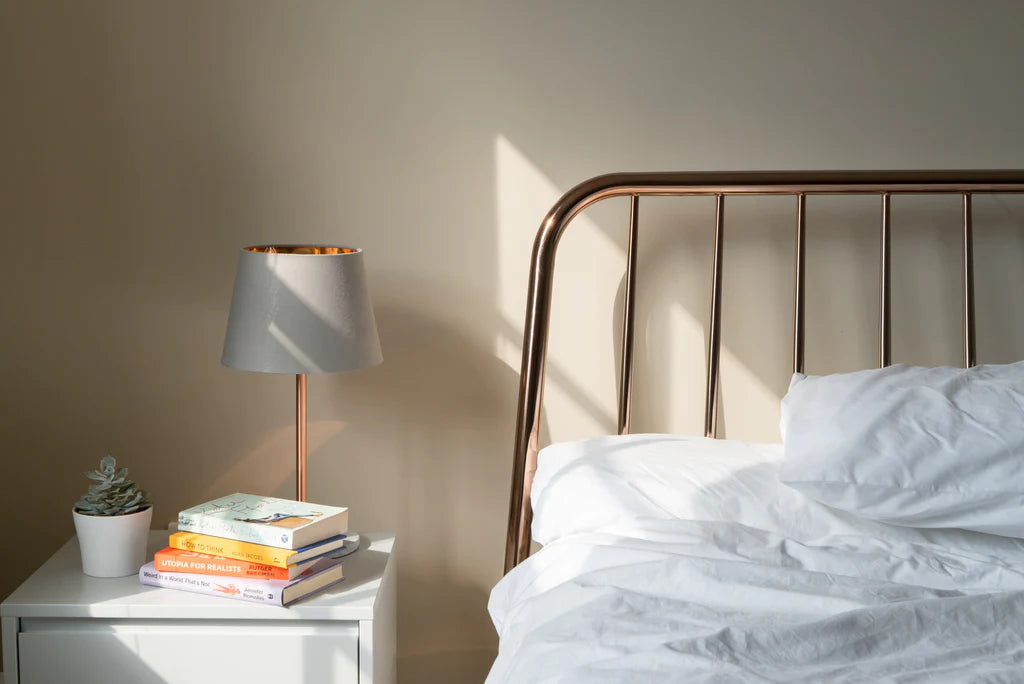 An elegant copper lamp with a grey lampshade sitting on a bedside table
