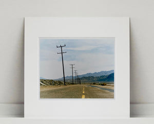 An open road in Amboy, California, featuring telephone poles disappearing into the mountainous distance. This classic and timeless landscape photograph is part of Richard Heeps' 'Dream in Colour' series. 