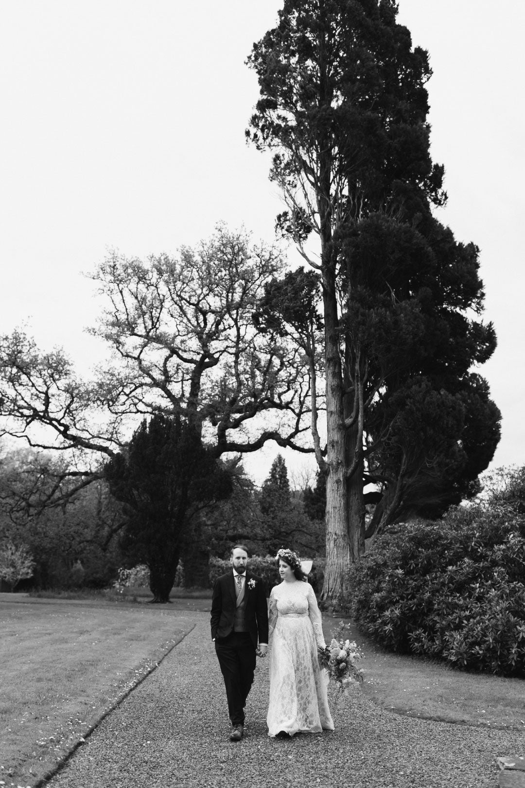 Black and white wedding photo of bride and groom walking with garden 