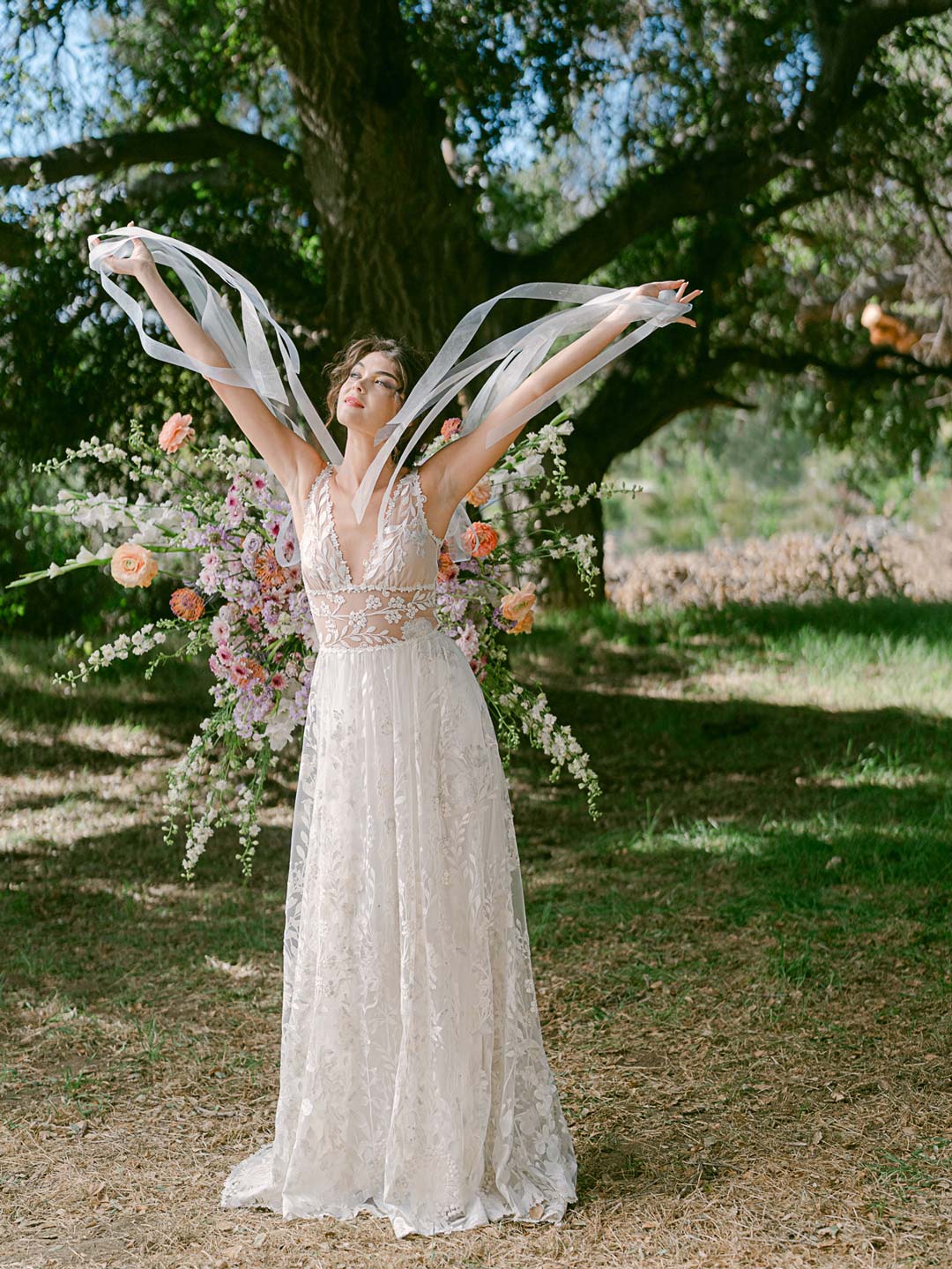 Bride in a lace wedding dress with flowing ribbons, standing in a garden under a large tree.