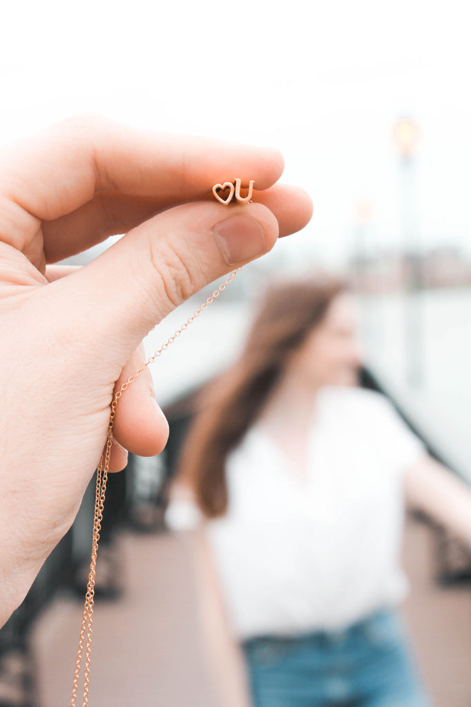 A girl stands near the water while a heart necklace is being held up