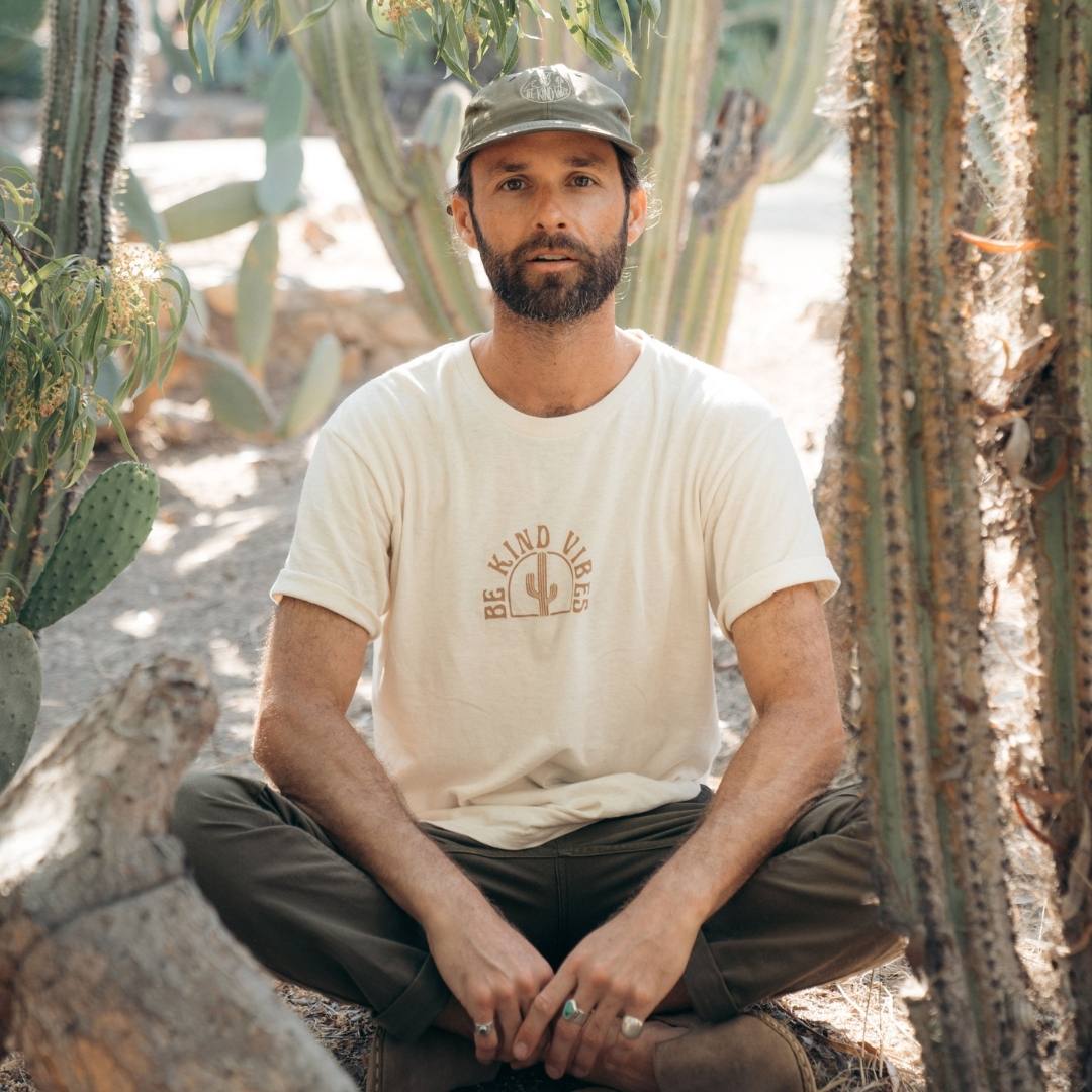 Image features a male model sitting crossed legged in a cactus garden wearing a green ball cap, green pants, and the Be Kind Vibes Desert Waves t-shirt.