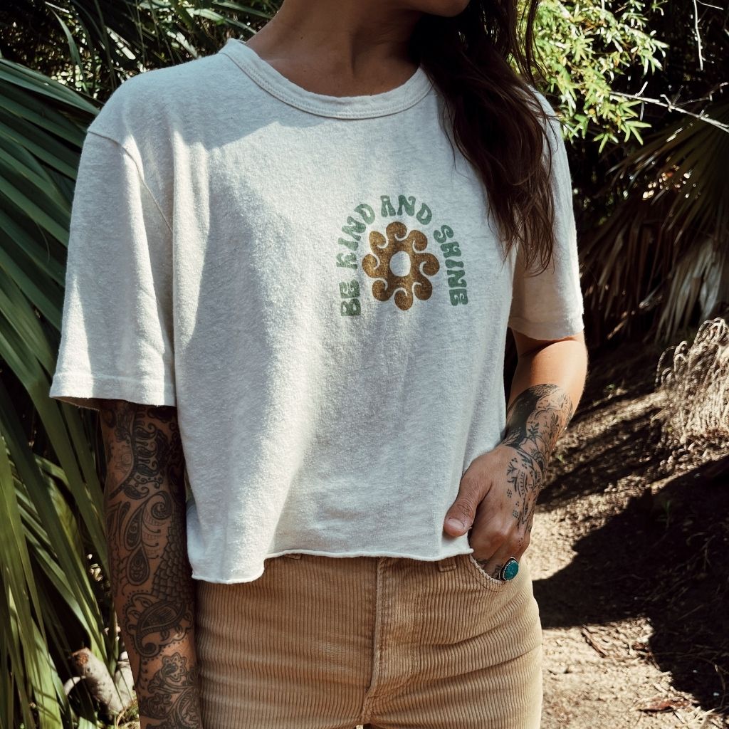 Hero image featuring a female model wearing the Be Kind Vibes Be Kind & Shine crop top in natural with khaki pants in a tropical setting.