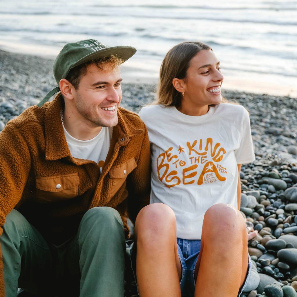 Image features two close friends sitting on the sand at the beach looking out at the horizon with a big smile on their faces. The male is wearing a Be Kind Vibes Hemp Hat in cactus with a rust colored jacket and green pants. The female is wearing the Be Kind Vibes 100% organic cotton Be Kind to the Sea t-shirt in natural with jean shorts. They're just having a ball.