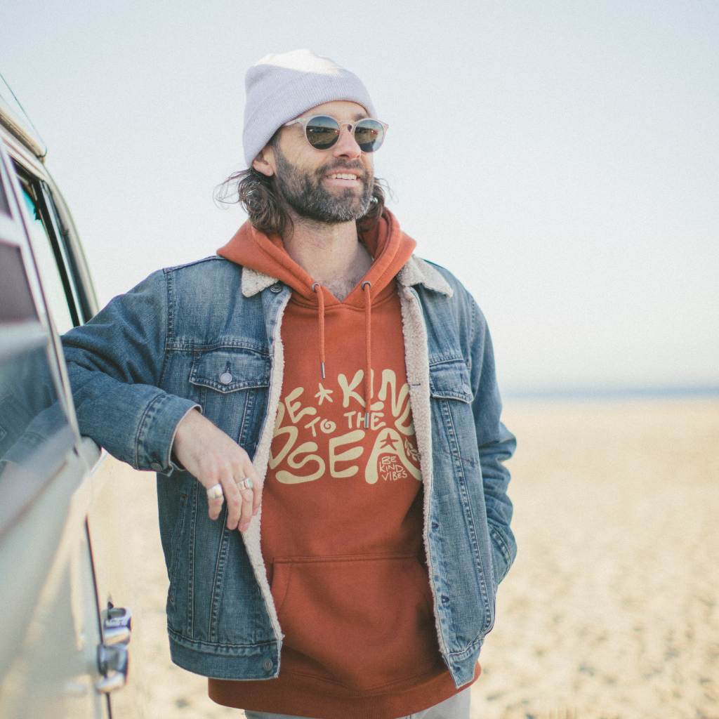 Image features a male model at the beach leaning against a VW bus wearing a grey touque, a jean jacket, and the Be Kind Vibes To the Sea hoodie.