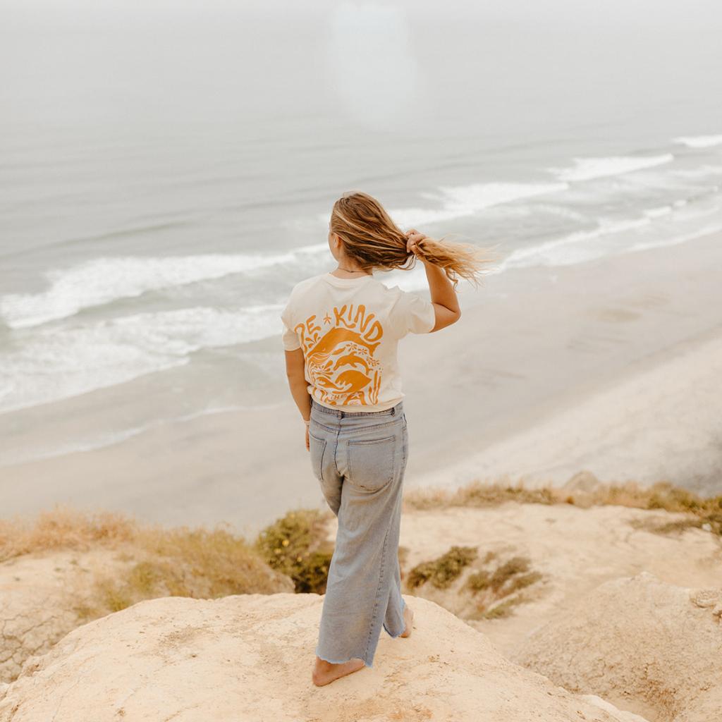Image features a female model standing on a cliff overlooking the ocean wearing the Be Kind Vibes organic To the sea t-shirt in natural with blue jeans.