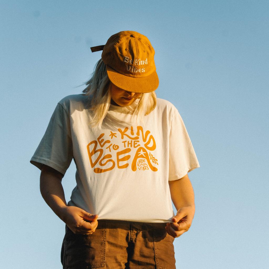 Image features a female model looking down at the t-shirt she's wearing which is the Be Kind Vibes organic To the Sea t-shirt in natural. She's also wearing the Be Kind Vibes Hemp Hat in rust as well as rust colored pants. The background is a bright blue sky with no clouds.