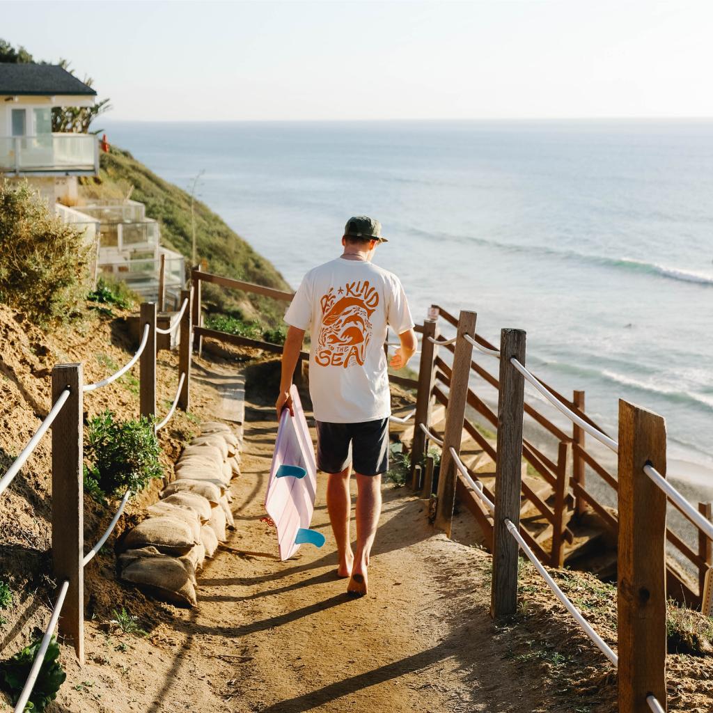 Image features a male model walking down a dirt path to the beach holding a surfboard and wearing the Be Kind Vibes organic To the Sea t-shirt in natural.