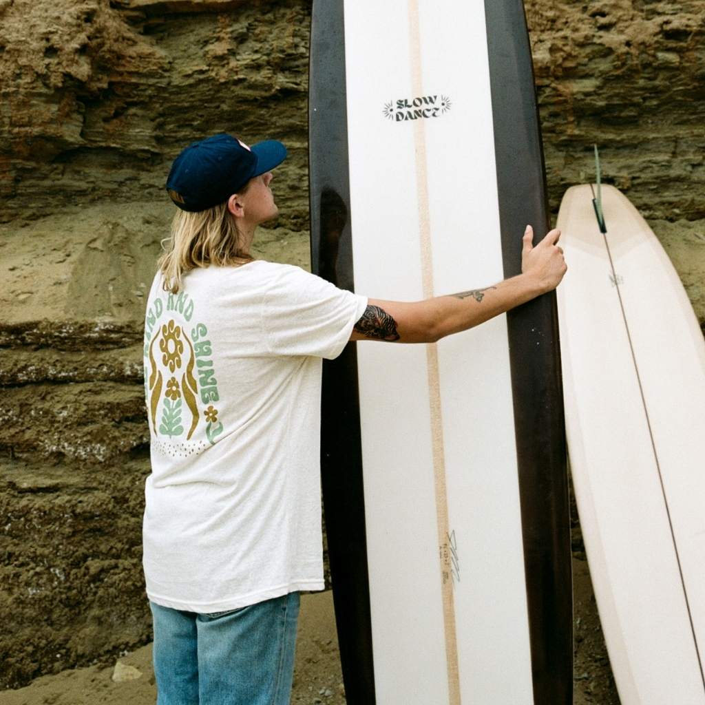 Hero image featuring the back of a male model holding a white and black surfboard wearing the Be Kind Vibes Be Kind & Shine tee in natural with blue jeans and a blue hat.