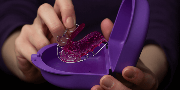 Image: An open purple retainer case with a person's hand holding a Hawley retainer.