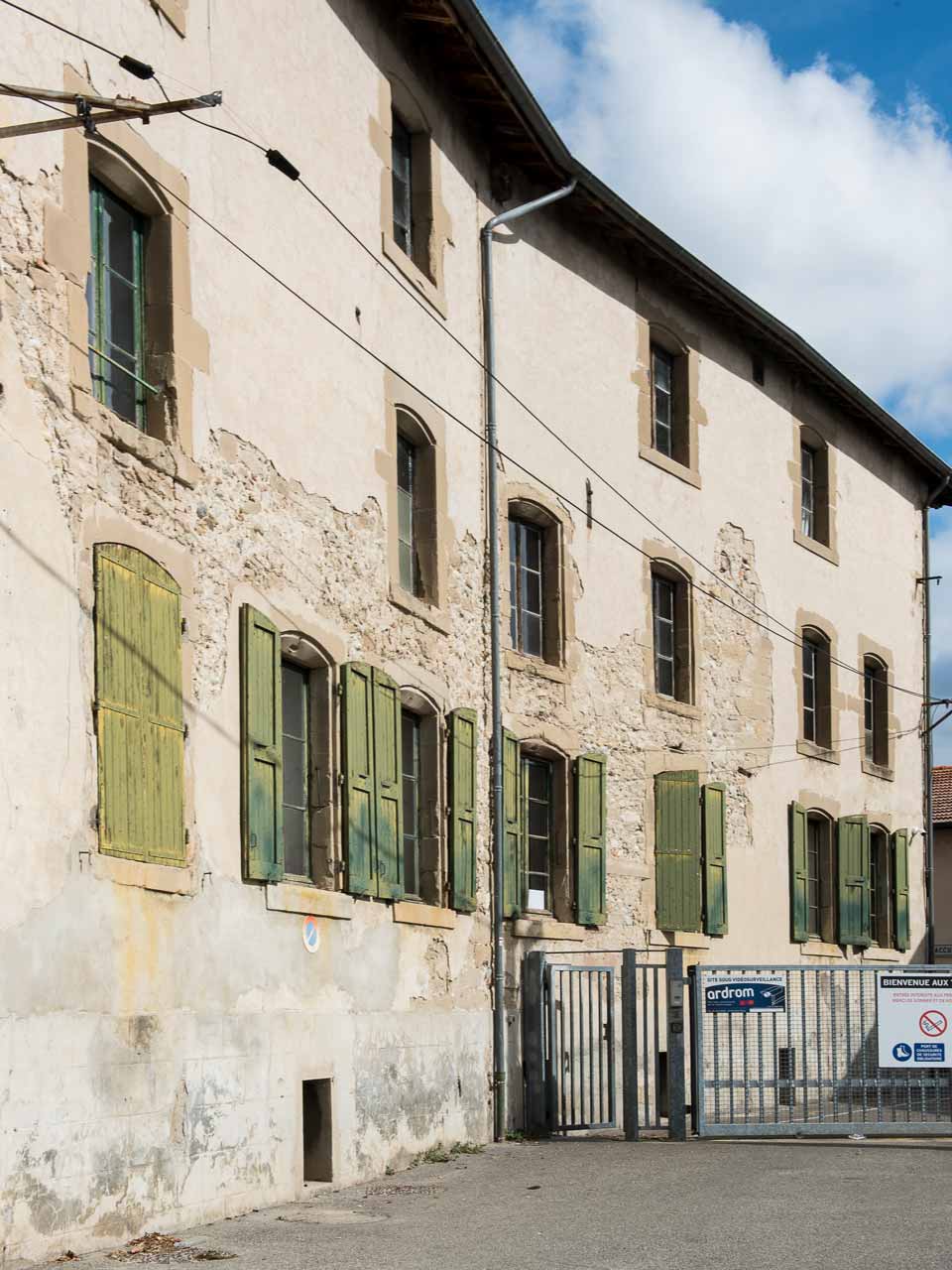 Historic facade of a French tannery building with weathered stone walls, green wooden shutters, and a gated entrance under a blue sky