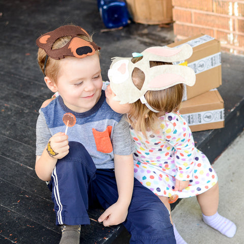 Little girl kissing brother next to their packaged gifts.  Wearing animal felt masks and eating lollipop candy.